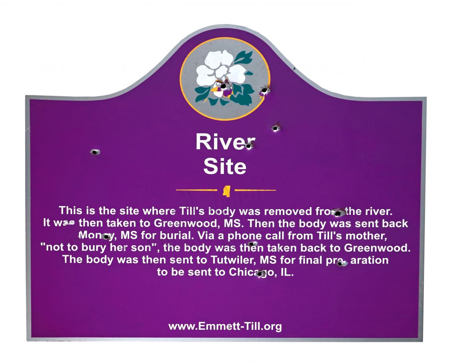 Purple sign with a white flower and the words "River Site" at top; it is riddled with several bullet holes