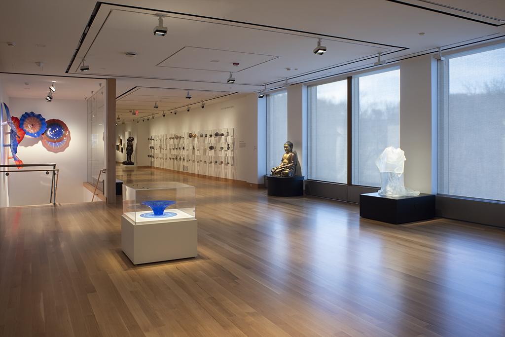 Photograph of a dimly lit art gallery with wooden floors and large windows; multiple sculptures line the walls