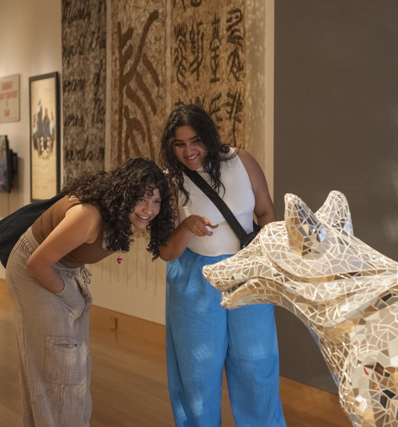 Two smiling young women with medium skin tone stand in an art gallery looking at a wolf sculpture covered in a mosaic of sparkling mirrors