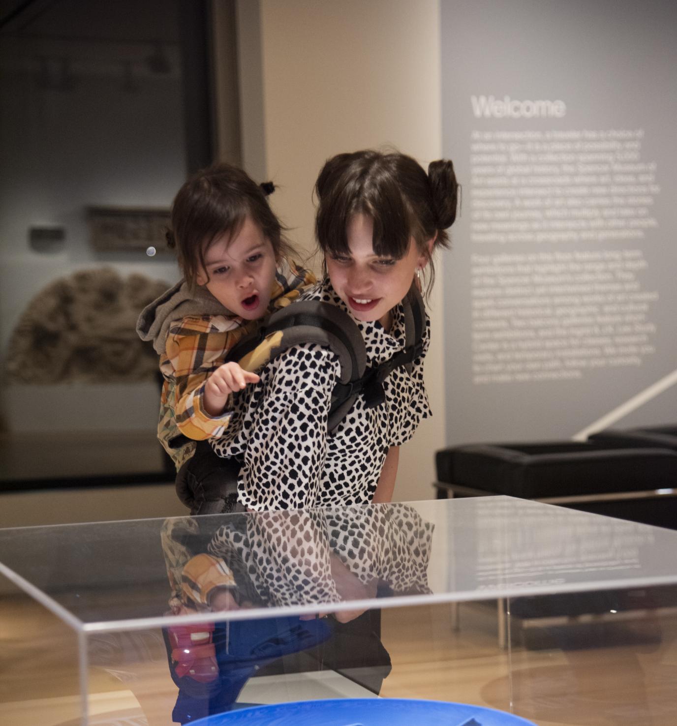 A young woman with dark hair carries a child on her back; both are looking at a blue glass sculpture in a gallery space