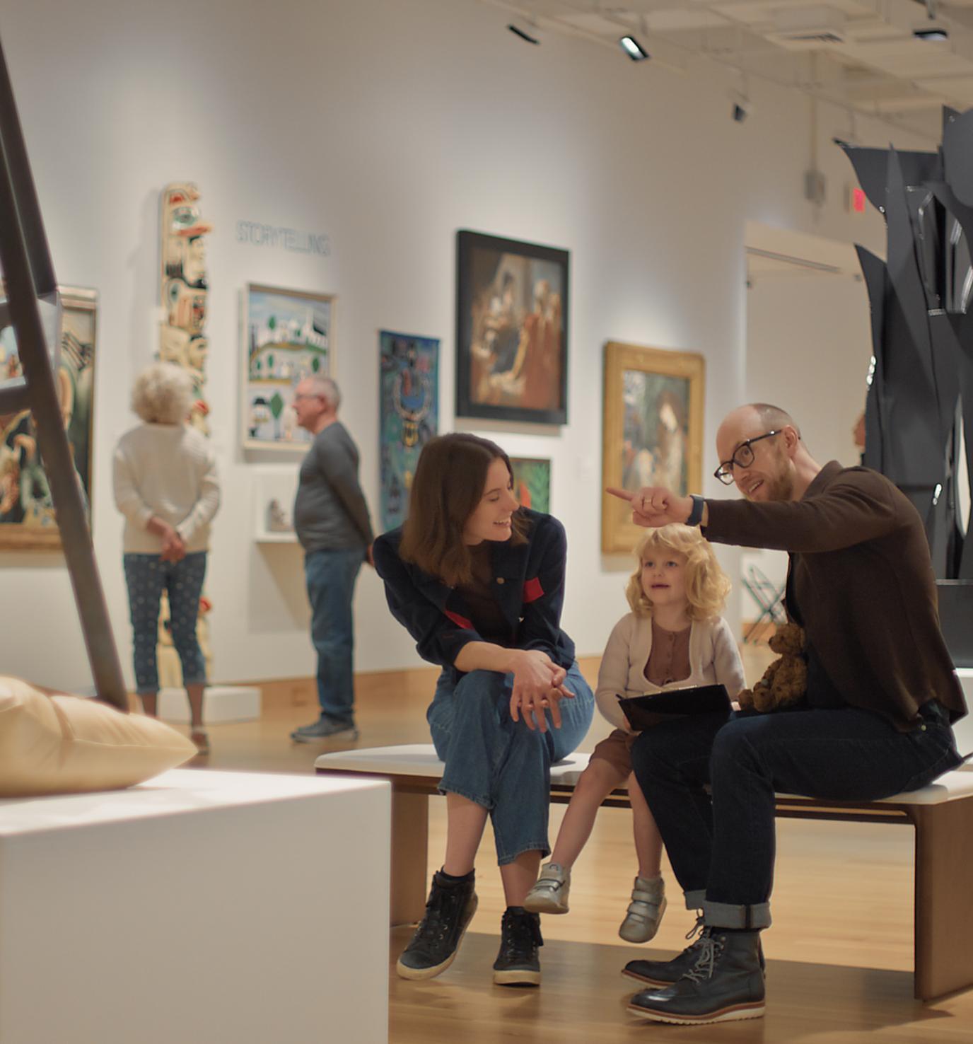 A woman, man, and young girl sit on a bench in an art gallery and look toward a sculpture of a chair