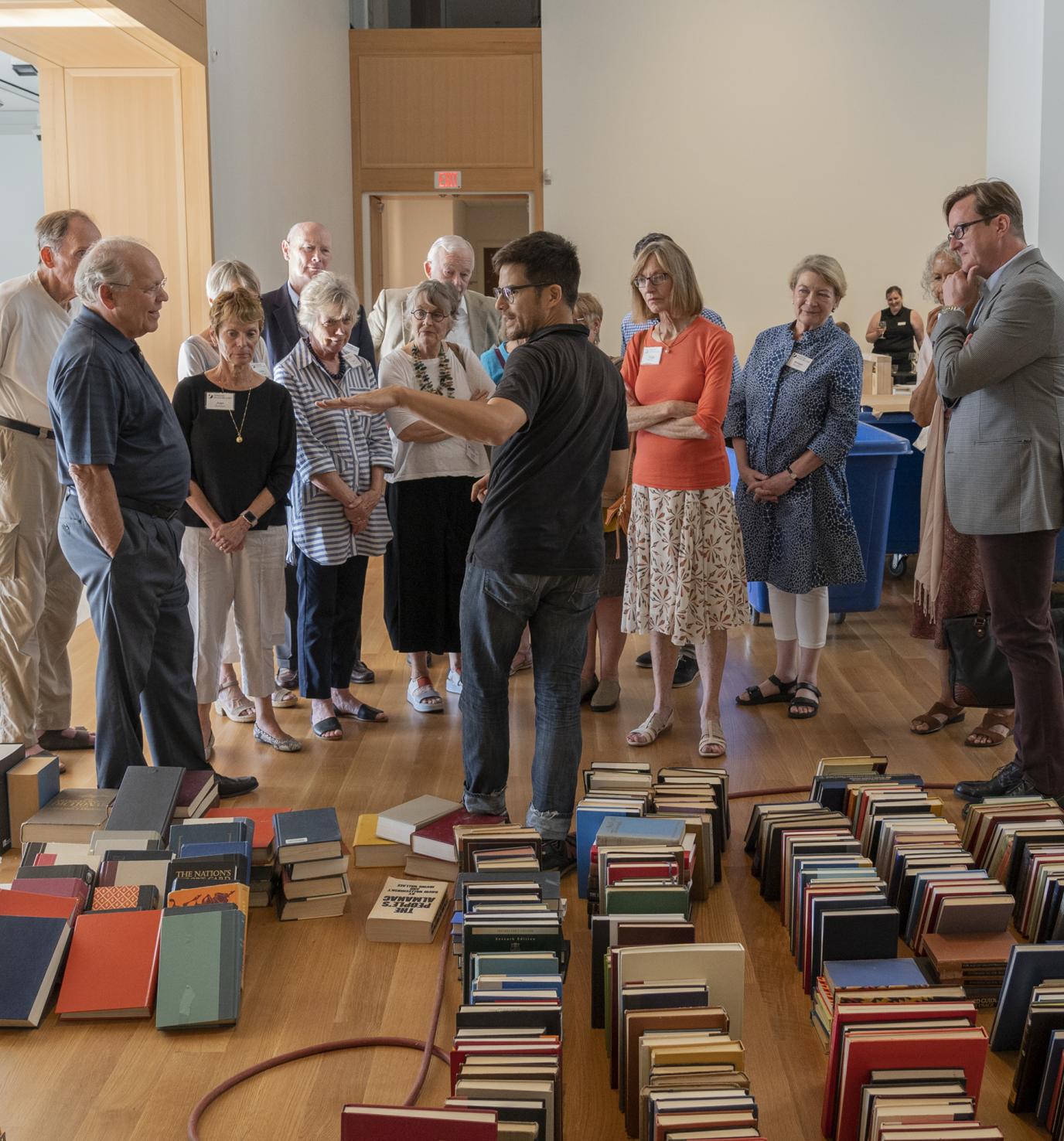 A man speaks to a crowd of people in front of stacks of books on a wood floor