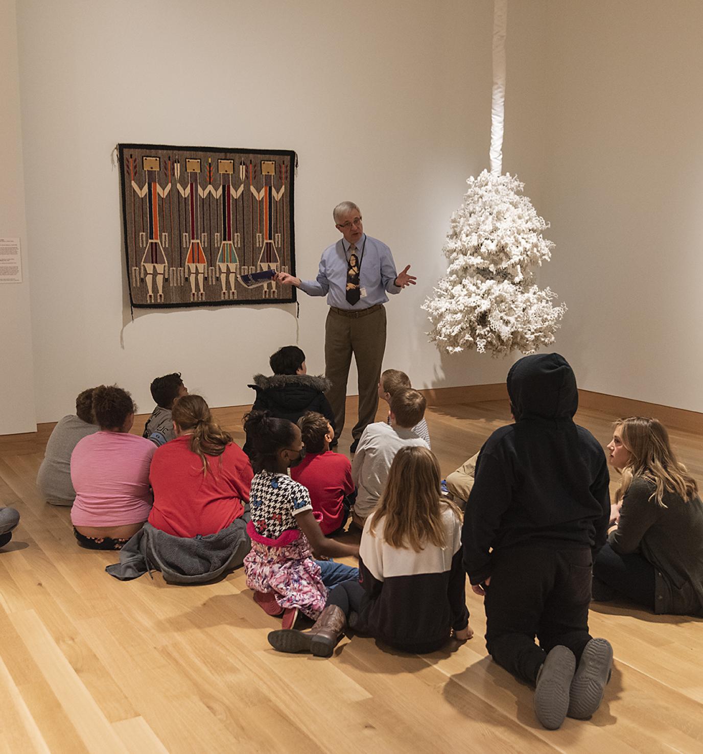 An older white man stands in front of a group of children seated in an art gallery