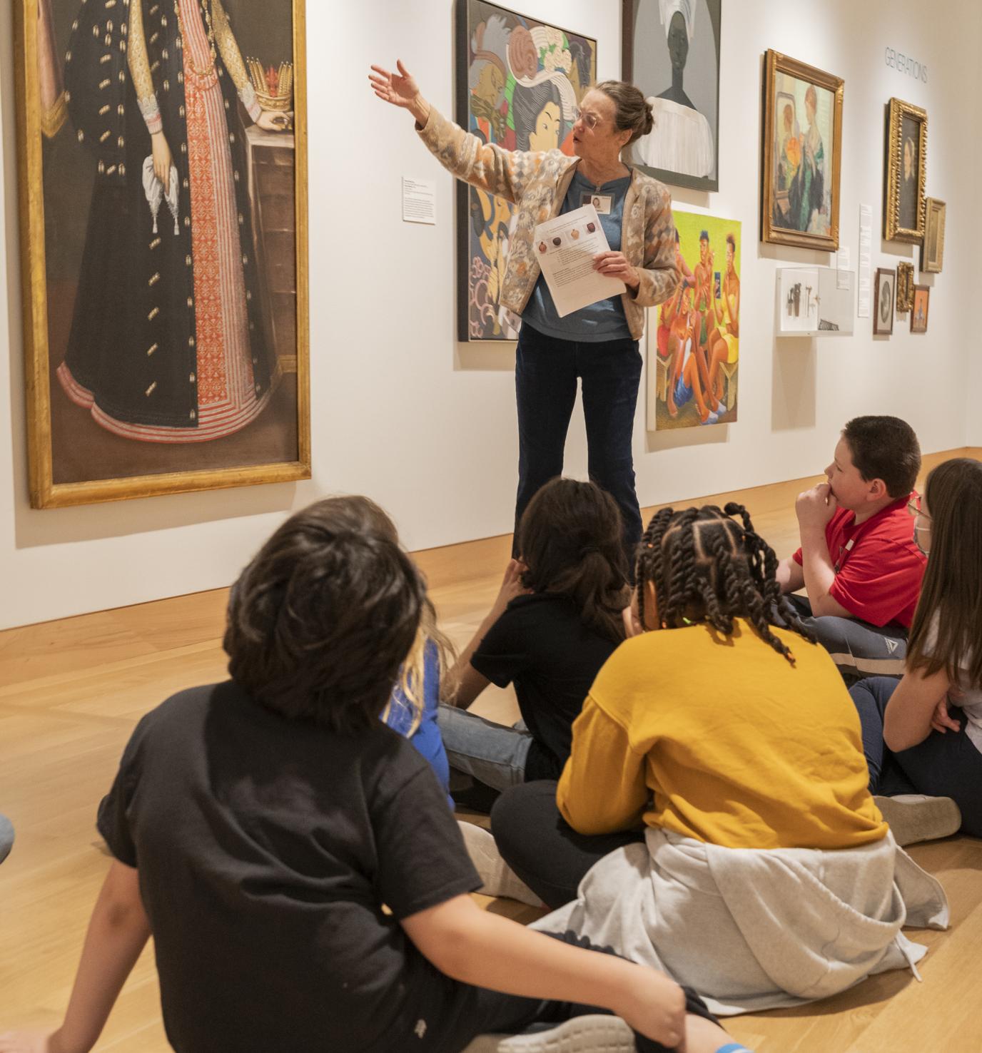 A woman stands in an art gallery in front of a group of seated children and gestures to a large painting of a royal woman on the wall