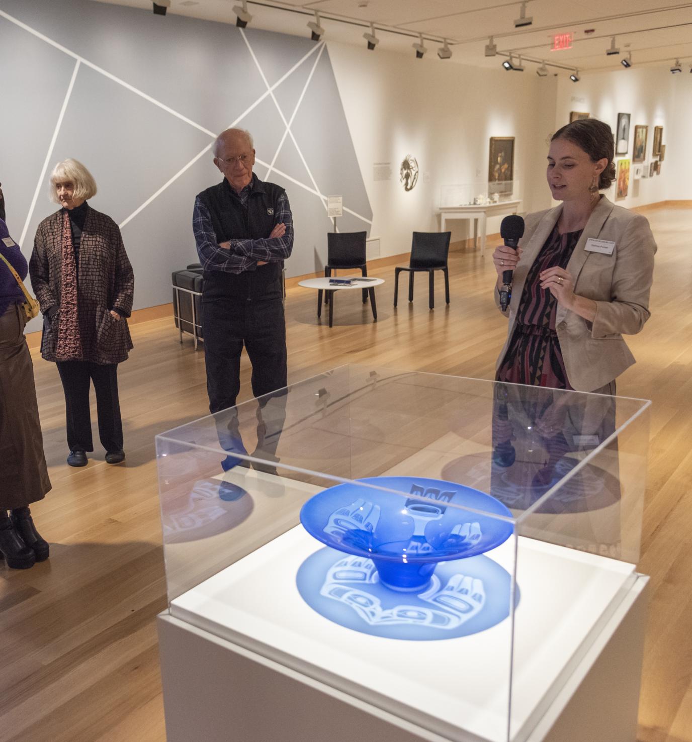 A white woman holding a microphone speaks to a group of people in a museum gallery