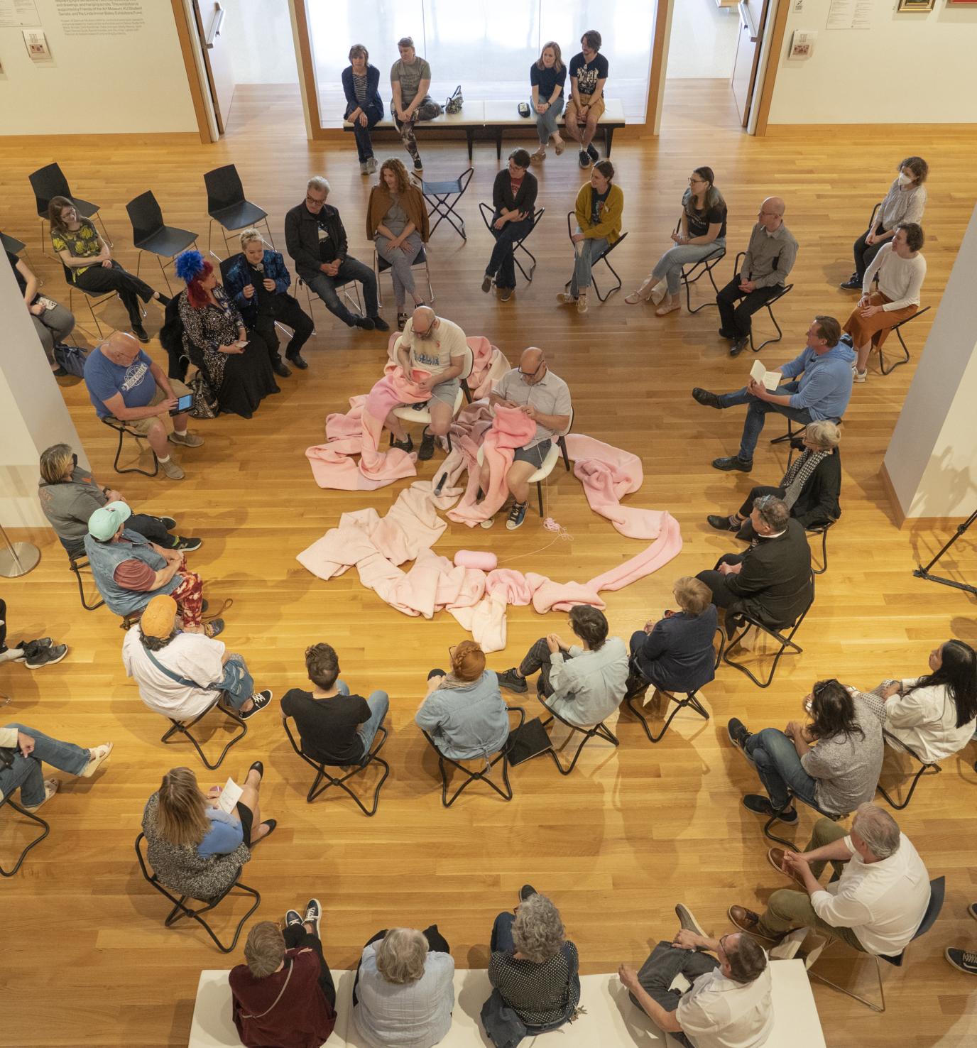 Two balding men crochet at either end of a long pink tube draped on the floor; a group of people sit in a circle around the men