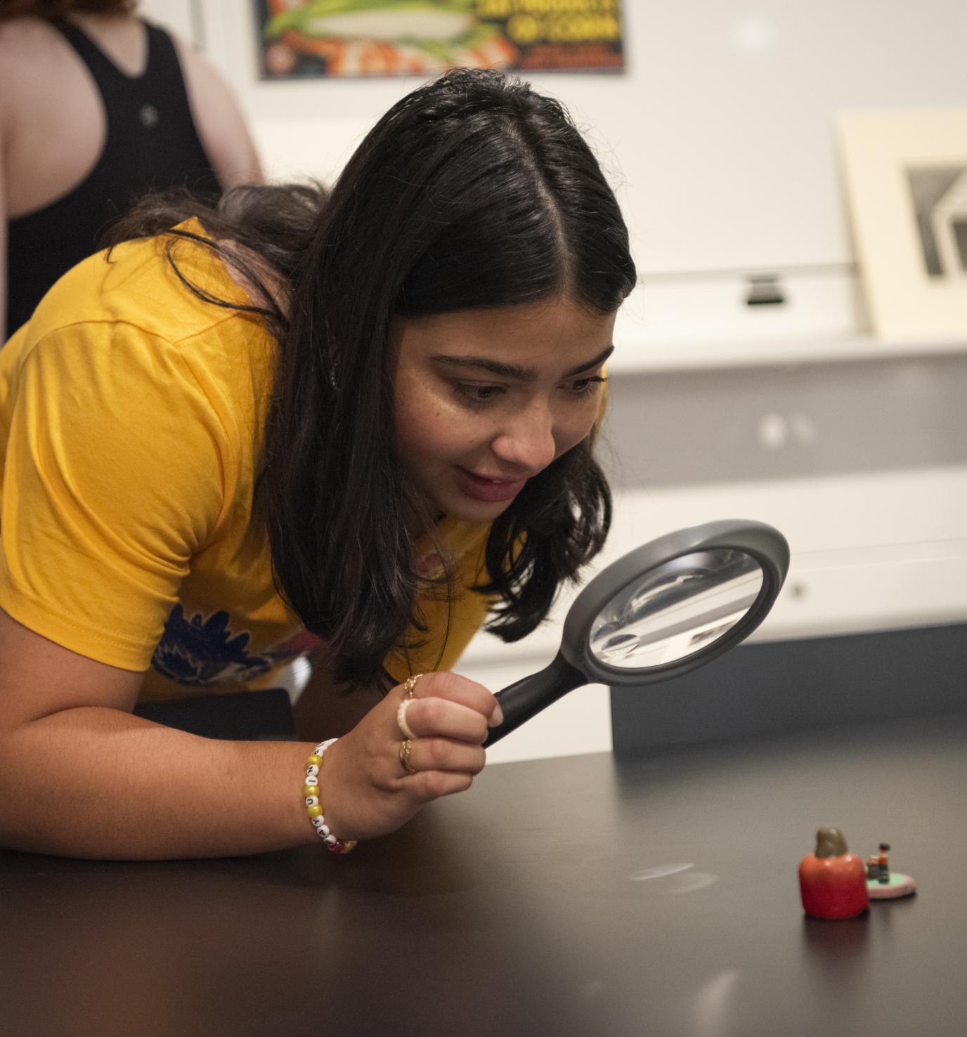 A young woman with brown skin and long dark hair leans over a table and holds a magnifying glass over a small object