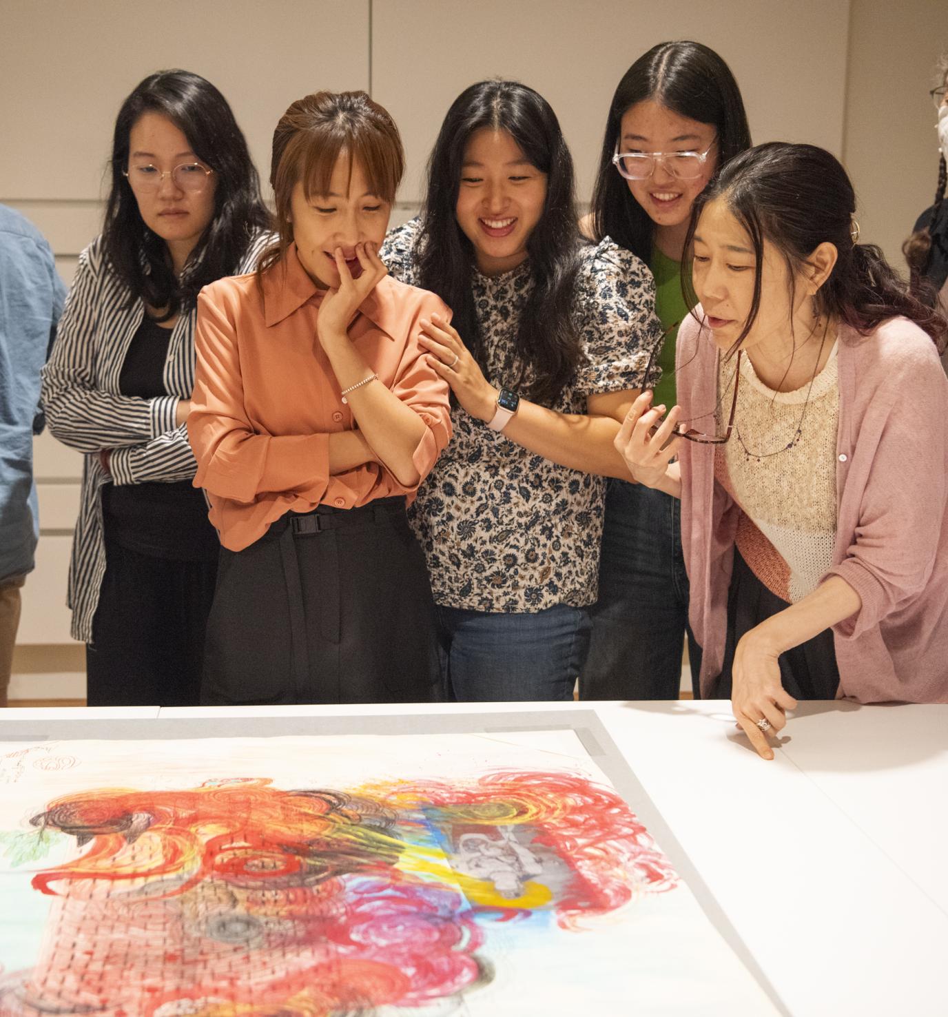 a group of Asian women gather around a colorful abstract print displayed on a white table
