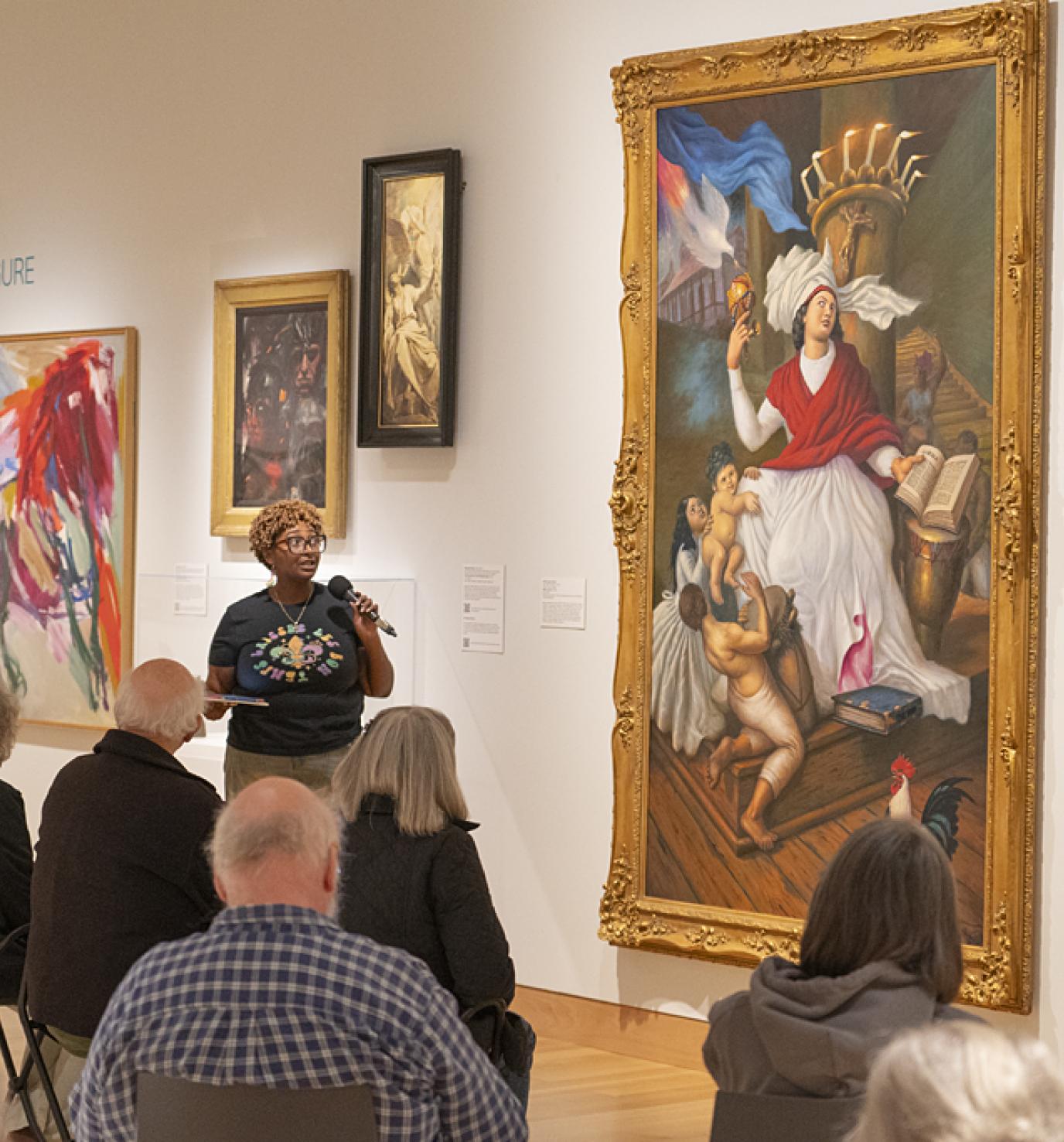 A young Black woman stands in front of a large painting and speaks into a microphone to a group of people