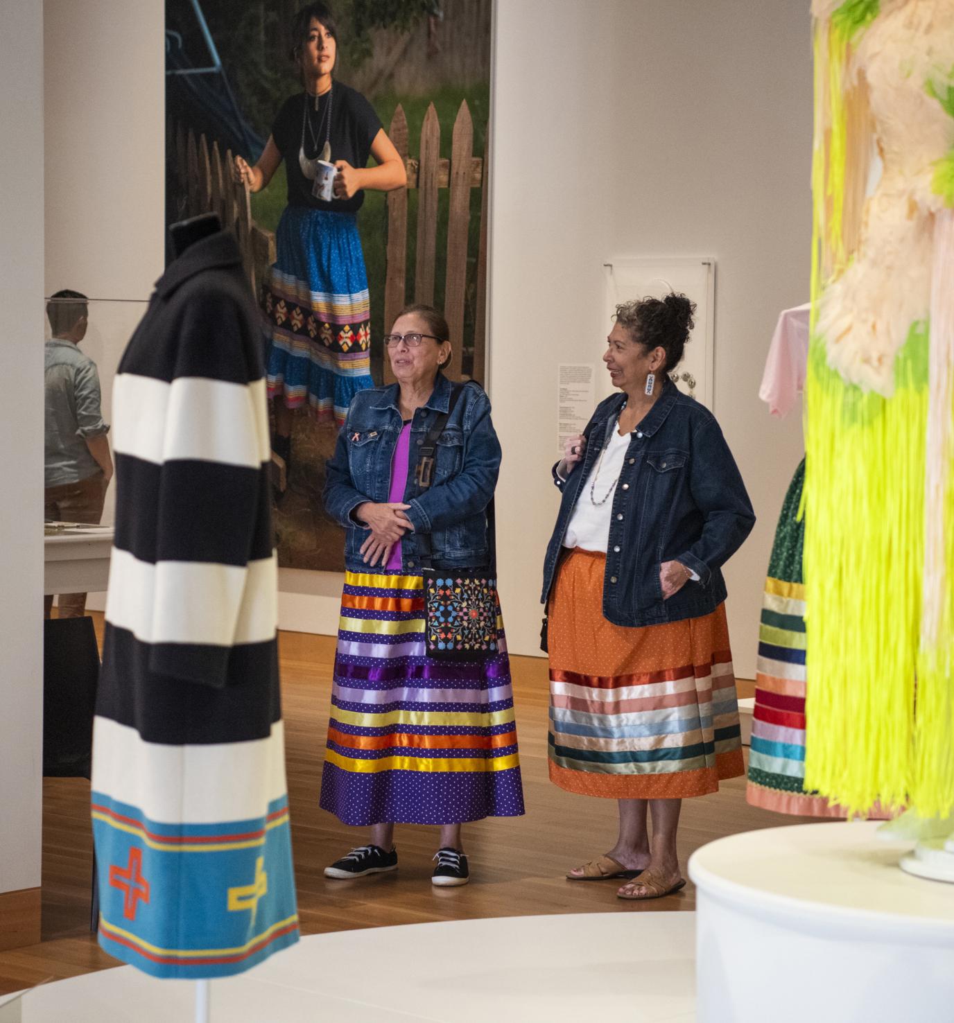 Two older Indigenous women wearing ribbon skirts stand in a gallery and look at artwork