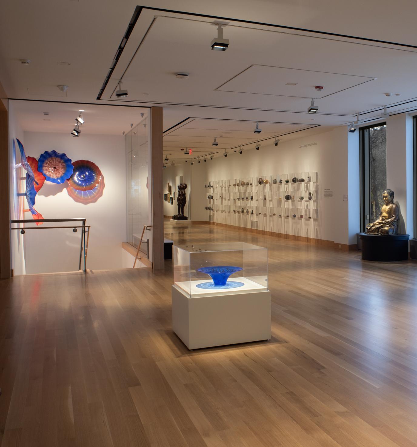 Photograph of a dimly lit art gallery with wooden floors and large windows; multiple sculptures line the walls