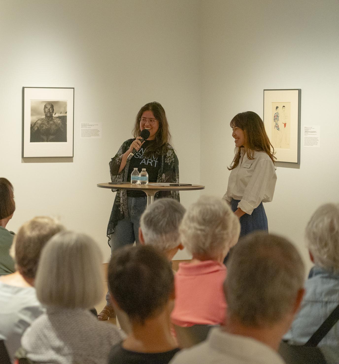 Two adults, one speaking into a microphone, stand in a gallery facing a group of seated people