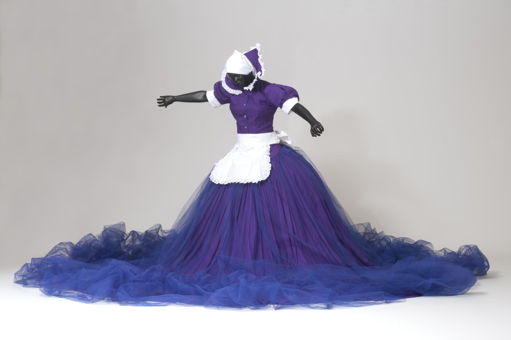 A Black female mannequin stands with outstretched arms, she wears a purple, blue, and white maid outfit with a large ballgown skirt 