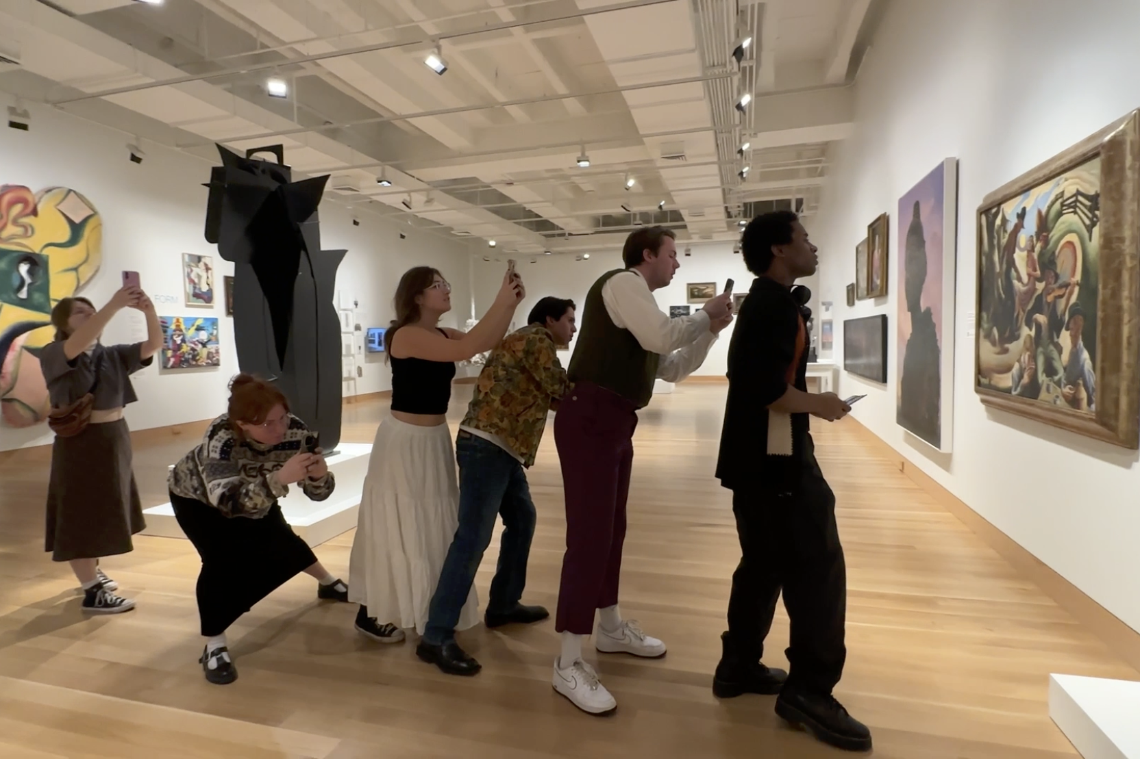 Six students stand in a single file line in an art gallery facing the same object and holding their cell phones in front of them