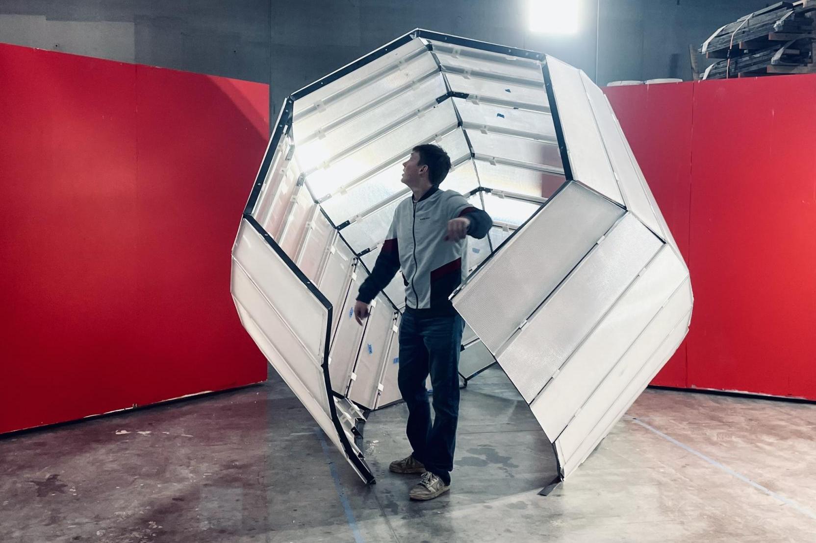 An adult stands inside a six-sided sculpture with an opening that rests on the ground of an industrial space