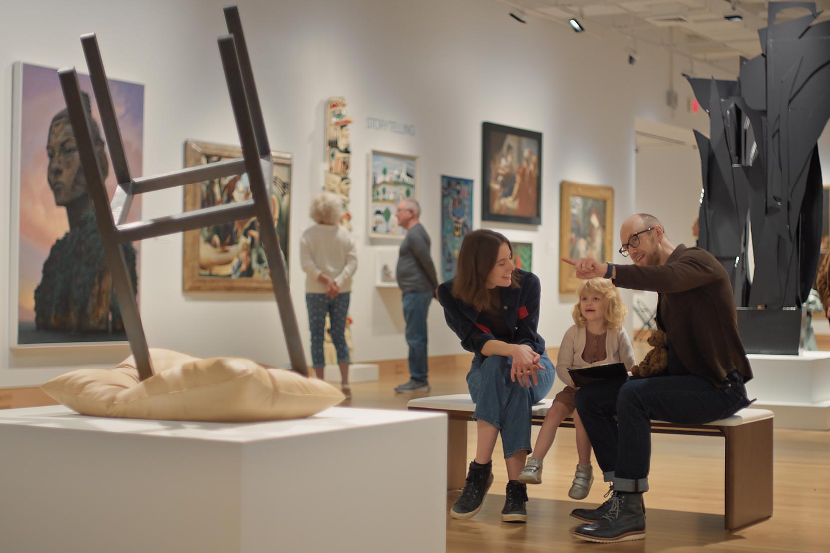 Two adults and a child sit on a bench looking at a sculpture in a museum gallery