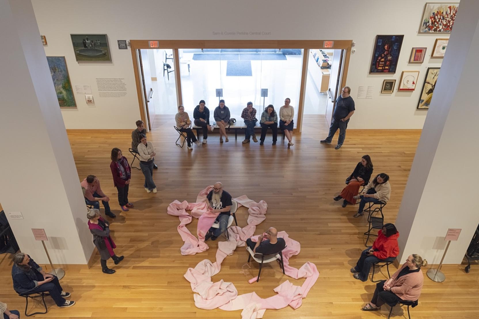 Two balding men sitting across from each other, both crochet at either end of a long pink tube draped on the floor 