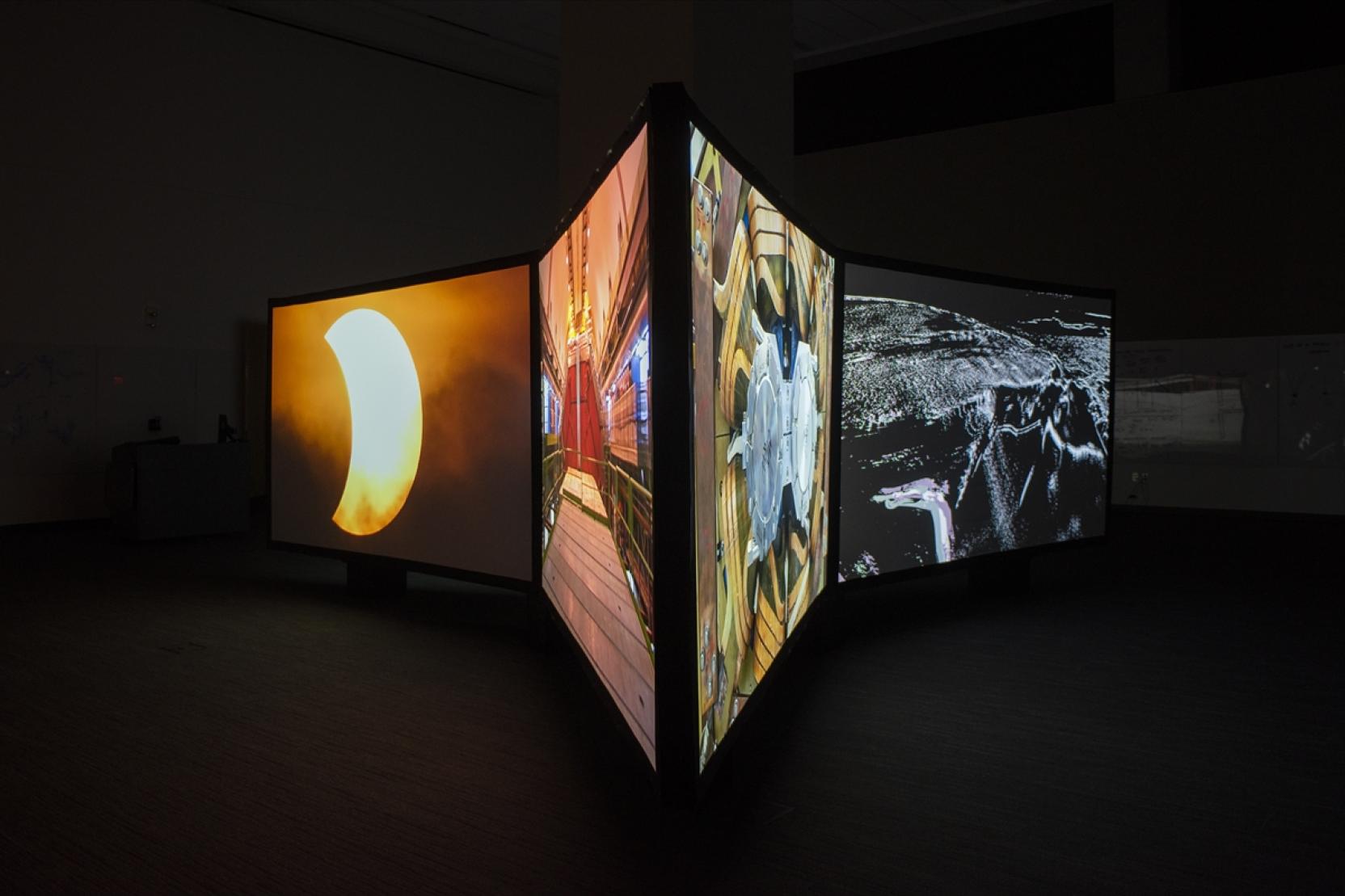 Four large screens in a dark room, each show a different colorful image; two face left and two face right 