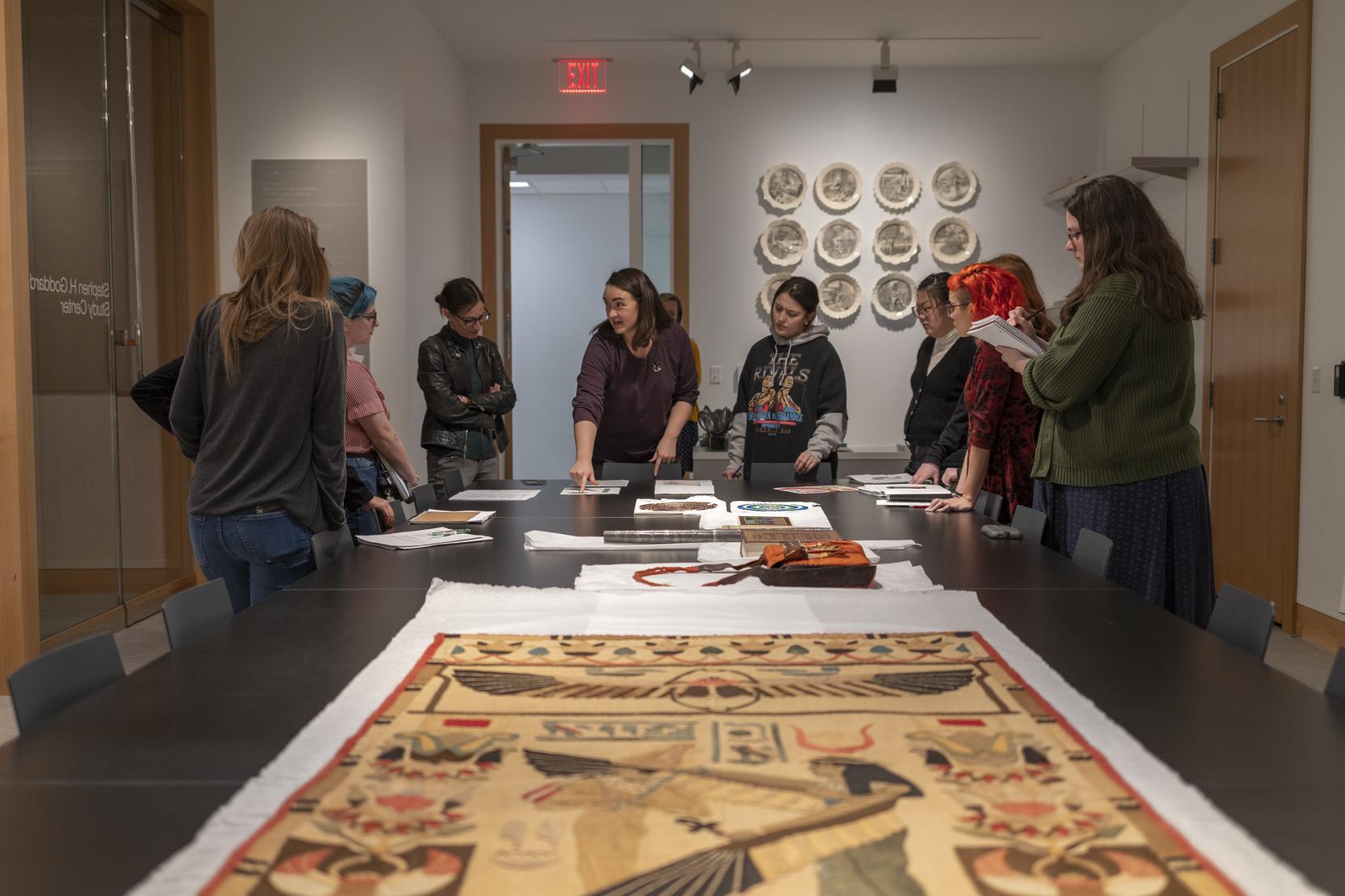 A white woman stands at the end of a large table covered in artworks with a group of young adults