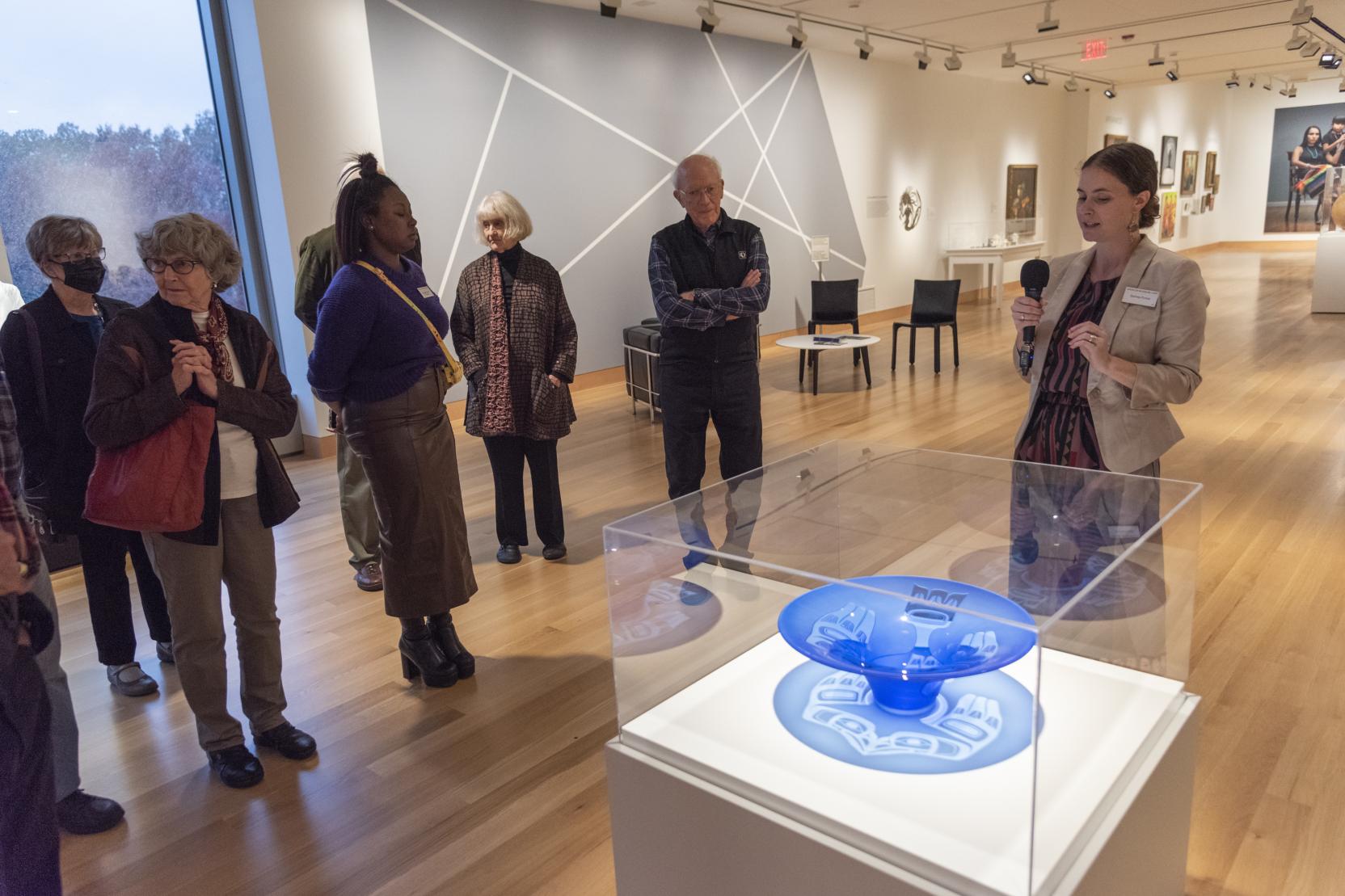 A woman with a microphone stands in an art gallery next to a blue bowl encased in glass; adults stand in front of her 
