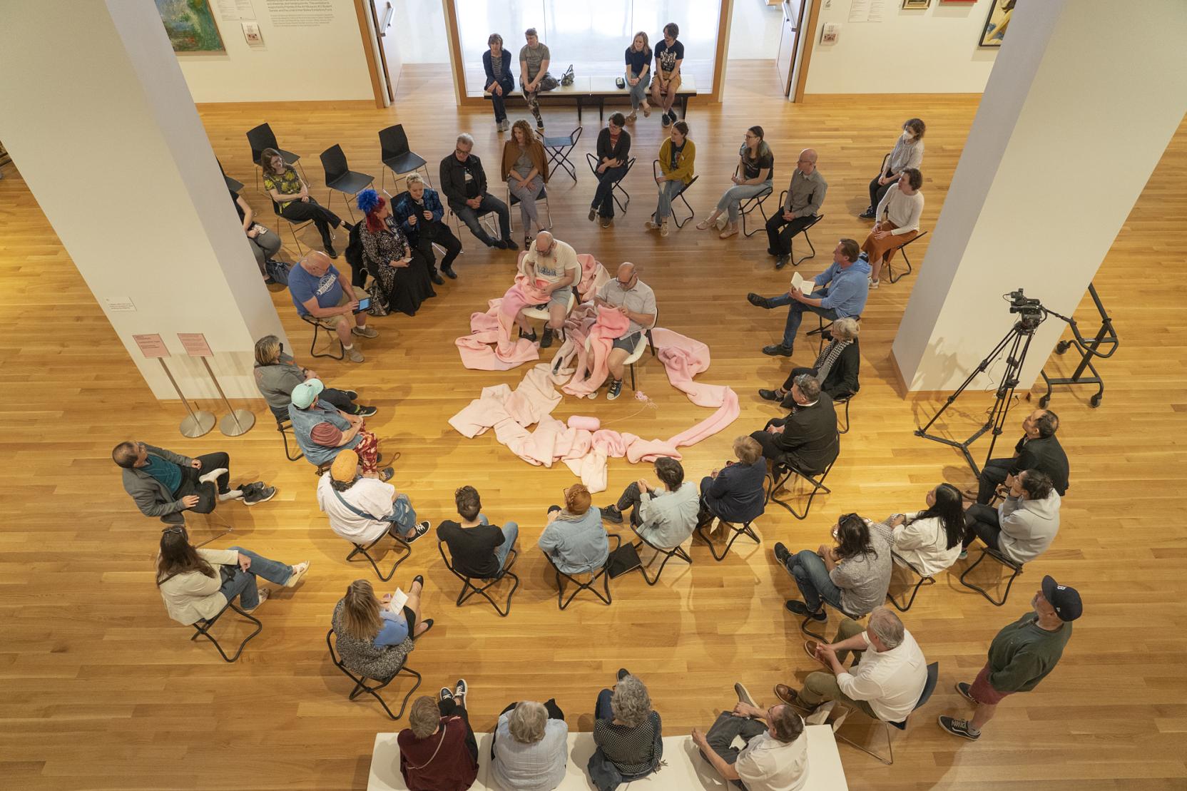 A group of people sit in a circle watching two men knit a large pink fabric piece