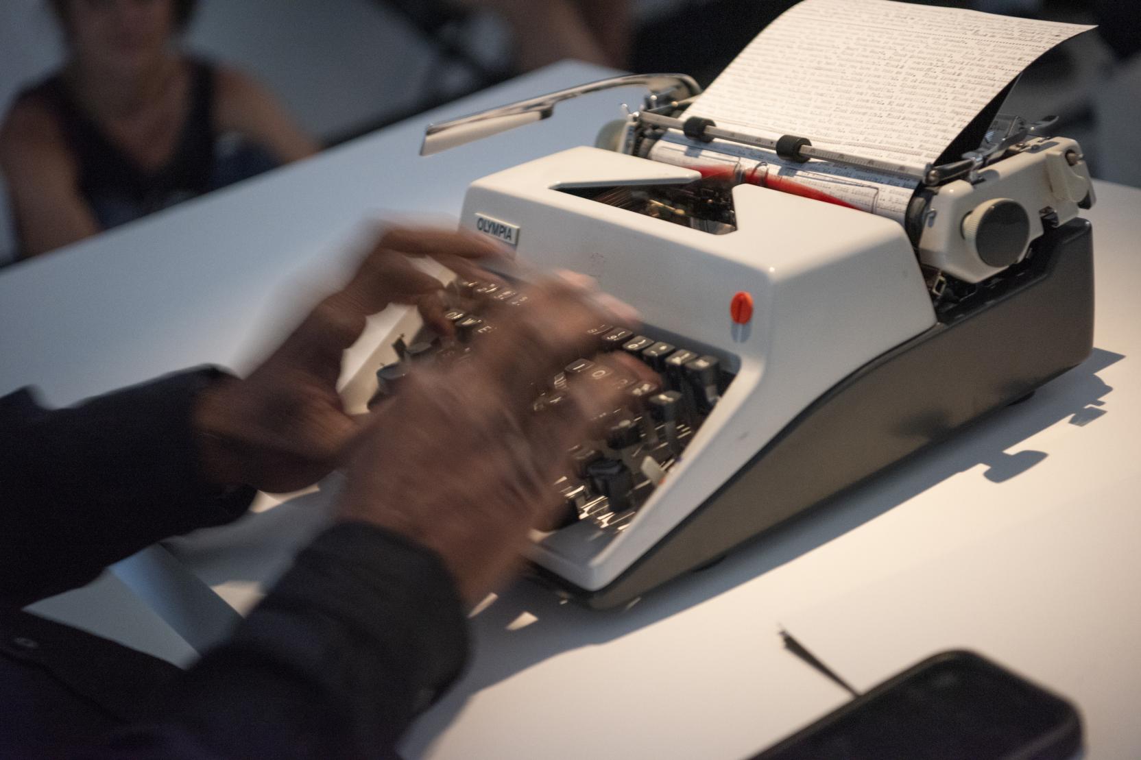 A Black person’s hands type at a small white typewriter, the hands appear blurry in the photo 