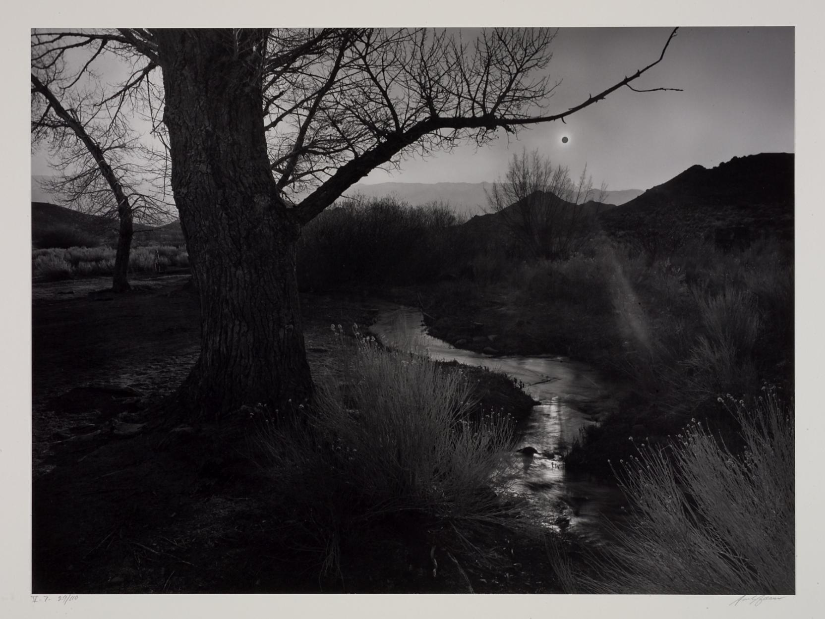 Black-and-white photo of trees, a stream, and hills; a pinpoint black sun is in the sky in the distance