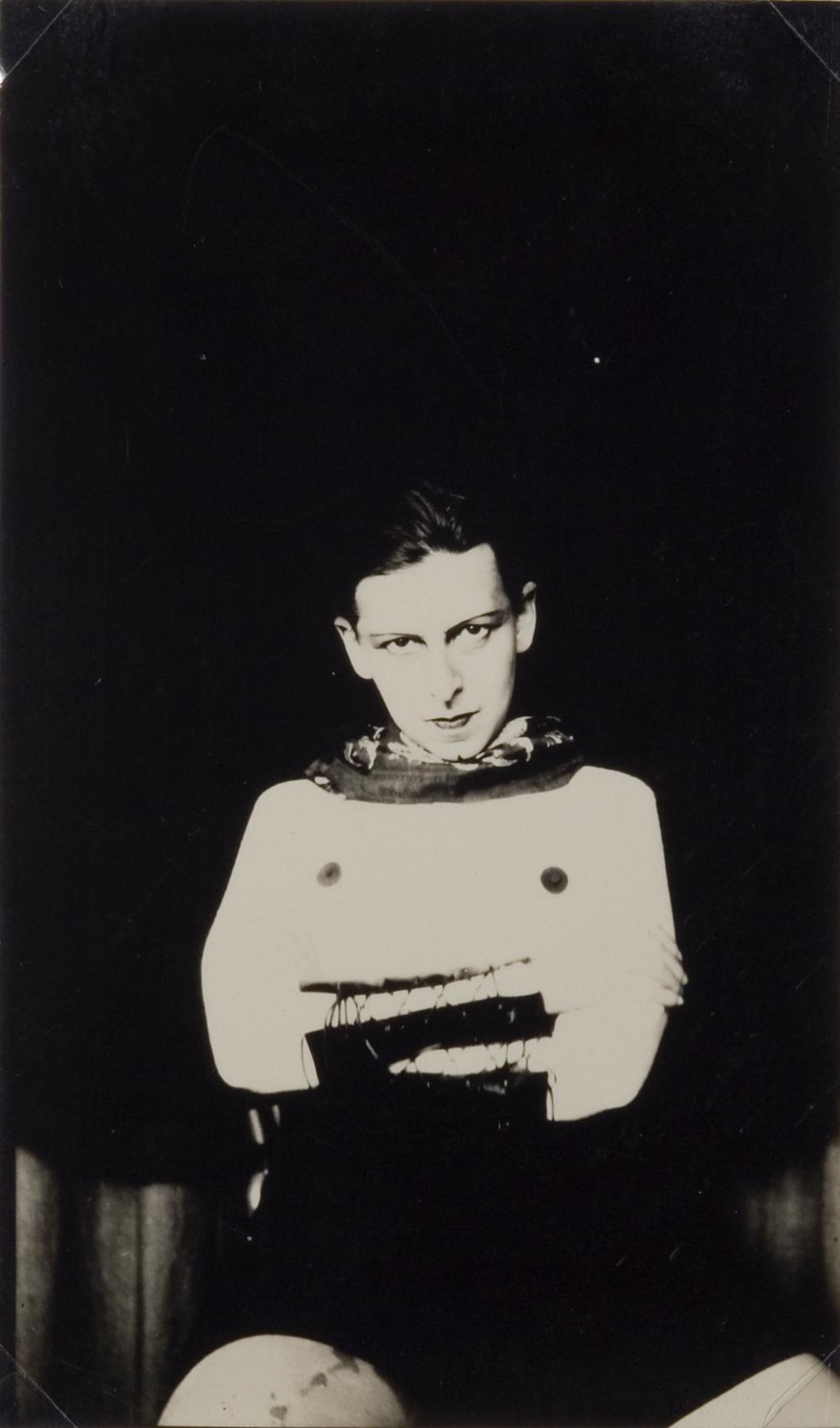 Black-and-white photo of a white adult sitting with their arms crossed staring definatly at the camera; they wear a scarf, stickers on their nipples, and black laced arm bands