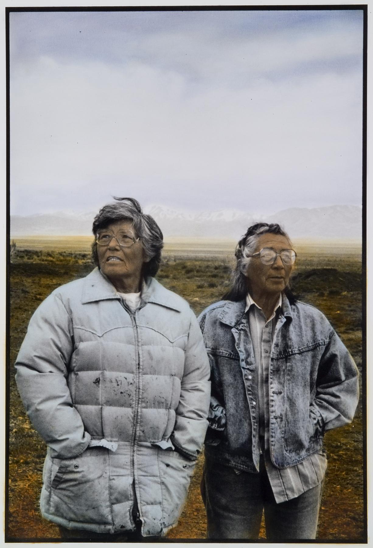Two elderly dark-skinned women wearing glasses and coats look away from the camera and stand in front of a mountainous landscape