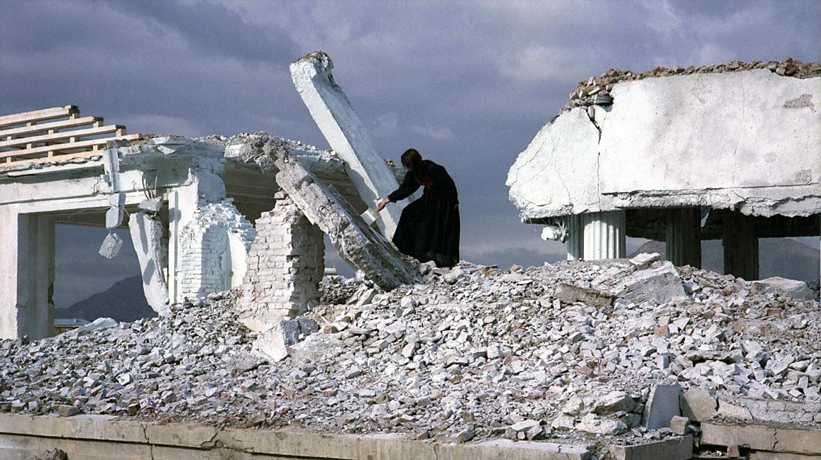 A figure dressed in all black stands in the rubble of a white building with her back to the camera