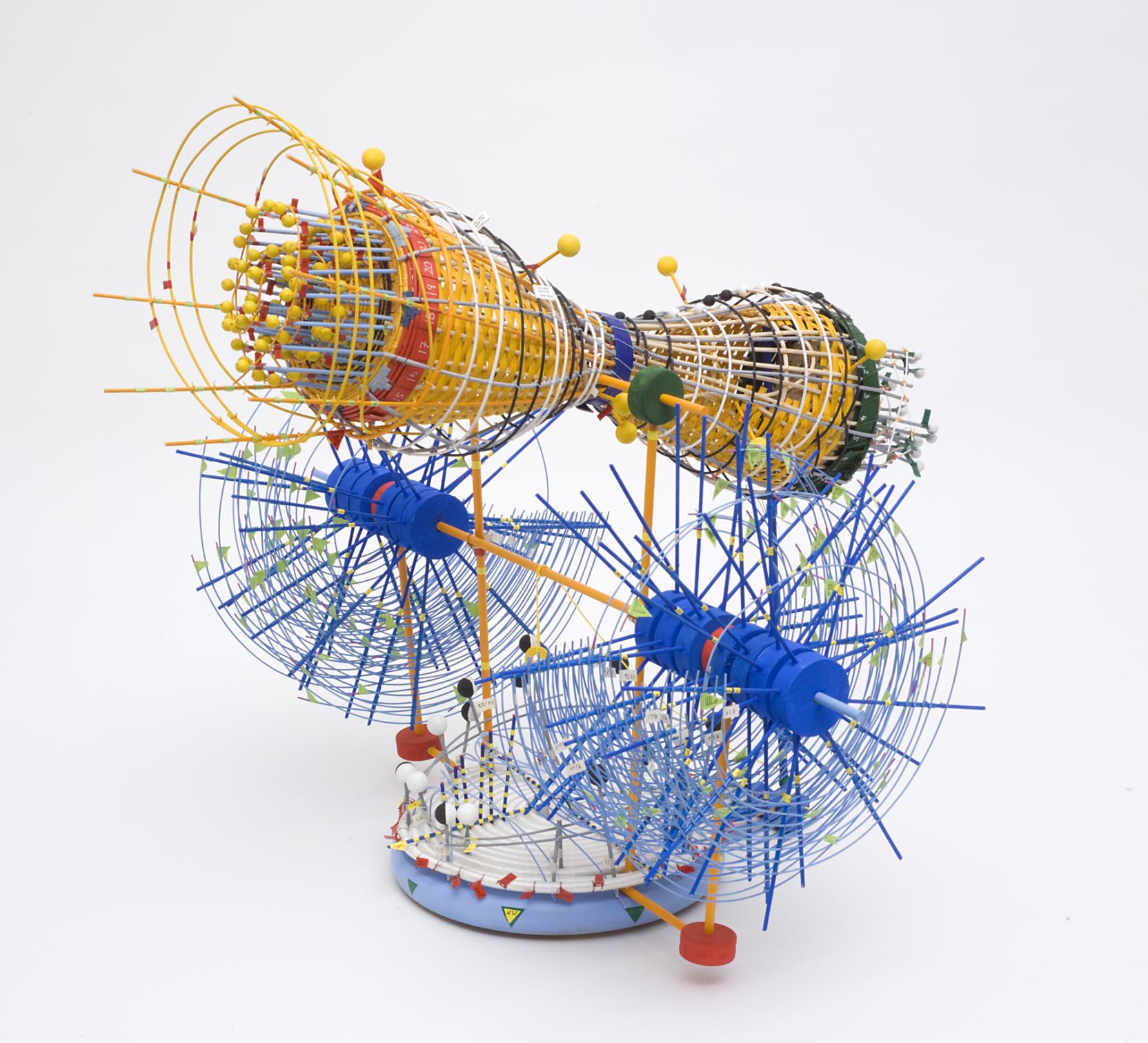 Abstract sculpture made of reeds woven together; a yellow hourglass shape rests above two blue wheels, both attached to a circular base covered small red flags