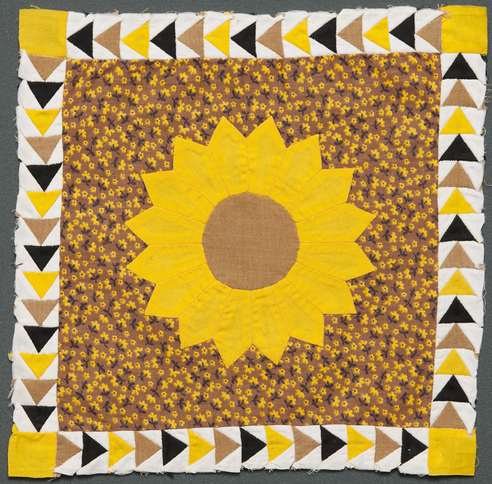 A quilt square with a yellow sunflower in the center of a floral-patterned background, bordered by white stripes with a sequence of yellow, brown, and black triangles