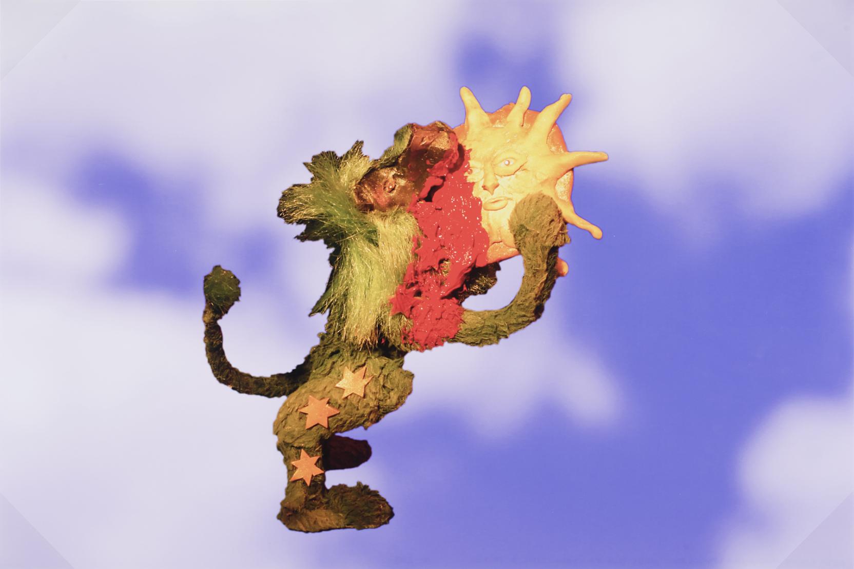 A green toy lion holds and eats yellow sun as blood runs down its face in front of a cloud-filled blue sky