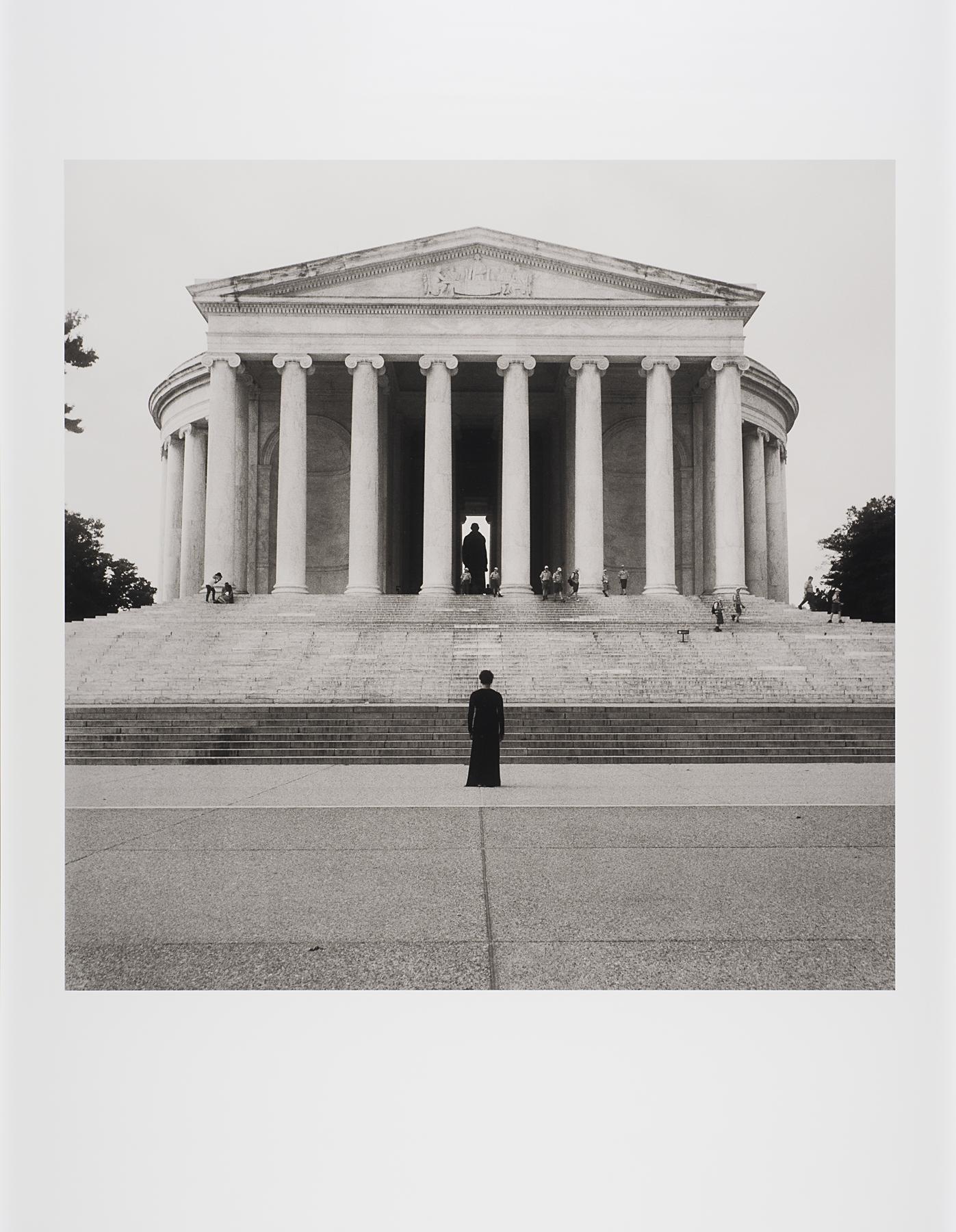 Black-and-white photograph of a figure in a long dark dress with their back to the camera standing below the steps leading to a large neoclassical building with columns