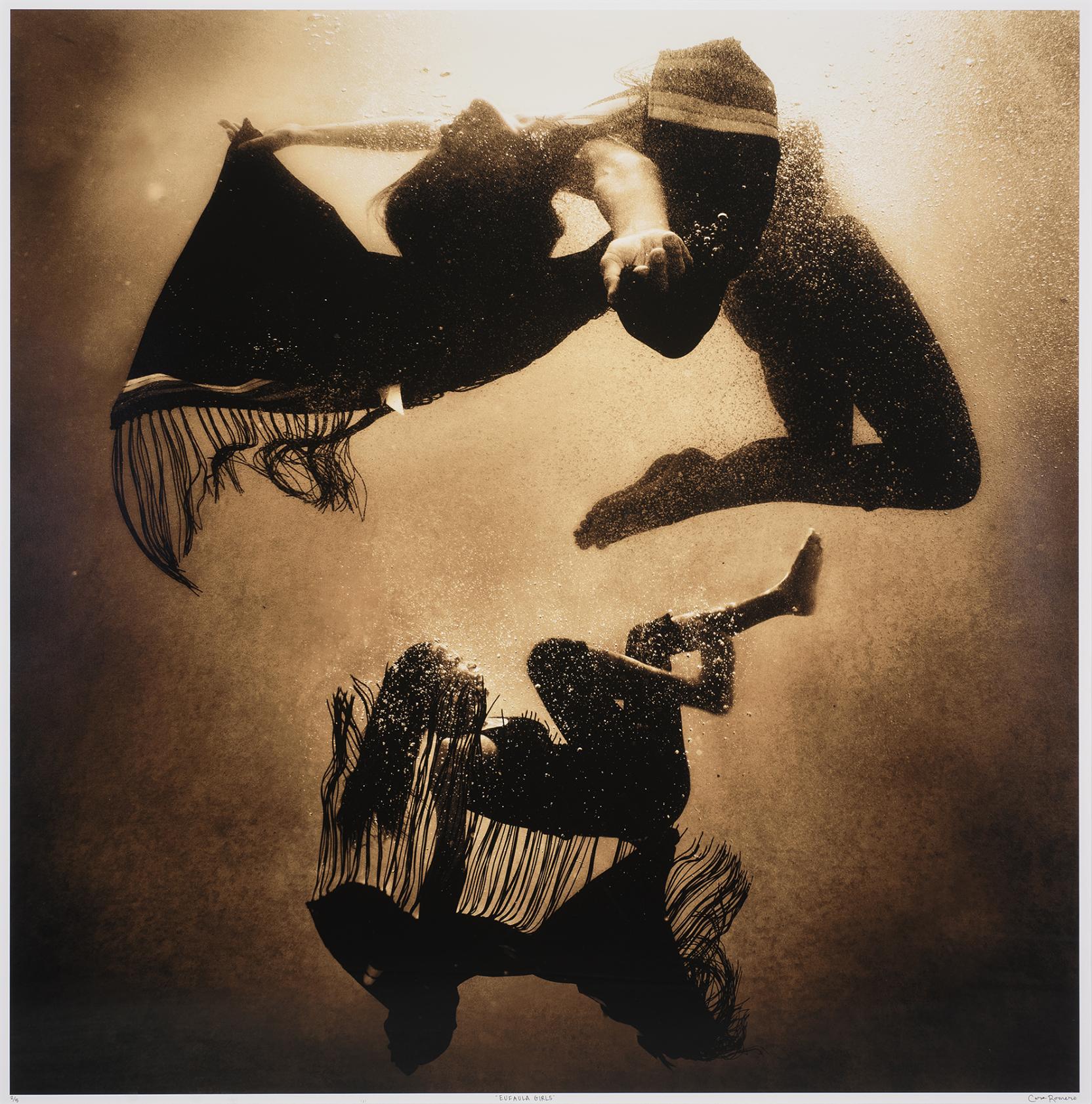 Brown-tinted photograph of two girls floating face up underwater with arms outstretched and fringed shawls flowing out behind them