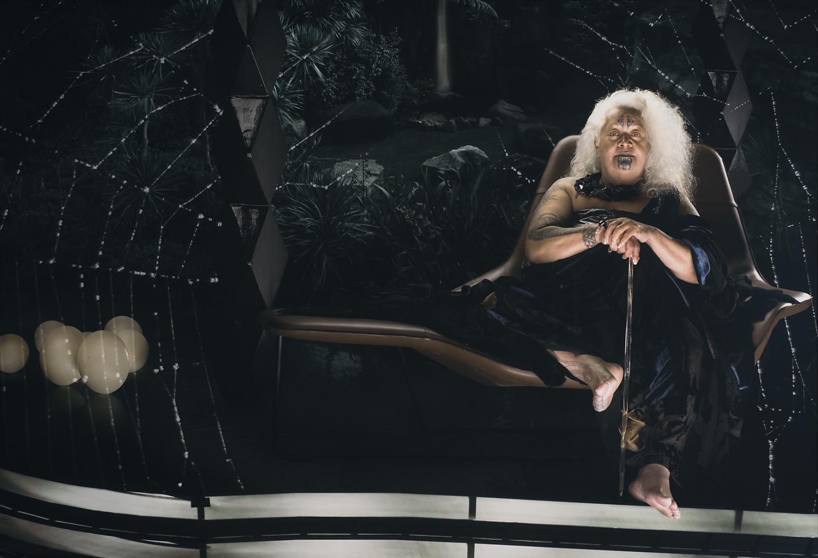 An old woman with white hair and facial tattoos sits holding a cane in a dark room surrounded by a large spiderweb 