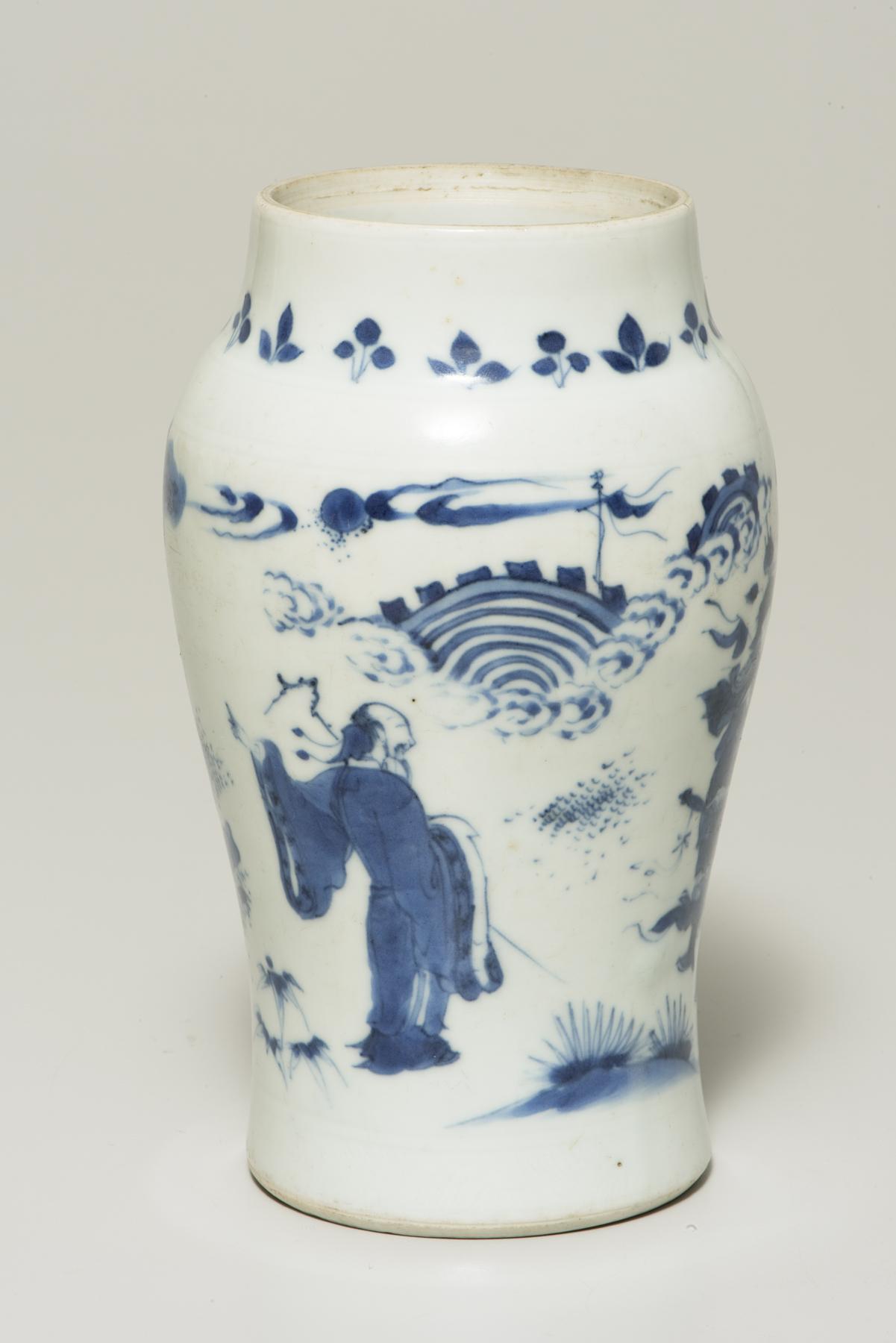 a tall white vase is decorated in blue with a warrior figure in the middle and a series of leaves around the top