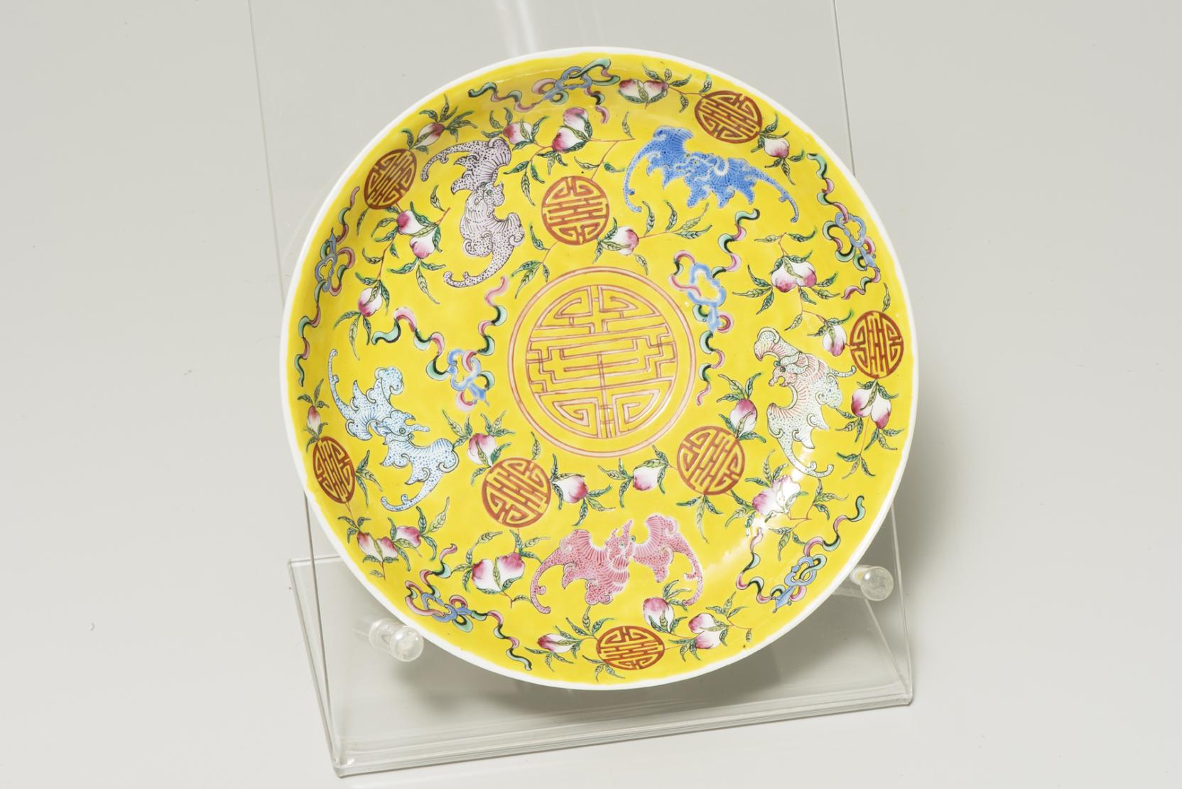 Yellow plate decorated with five bats in shades of blue and pink floating around a central pink medallion    