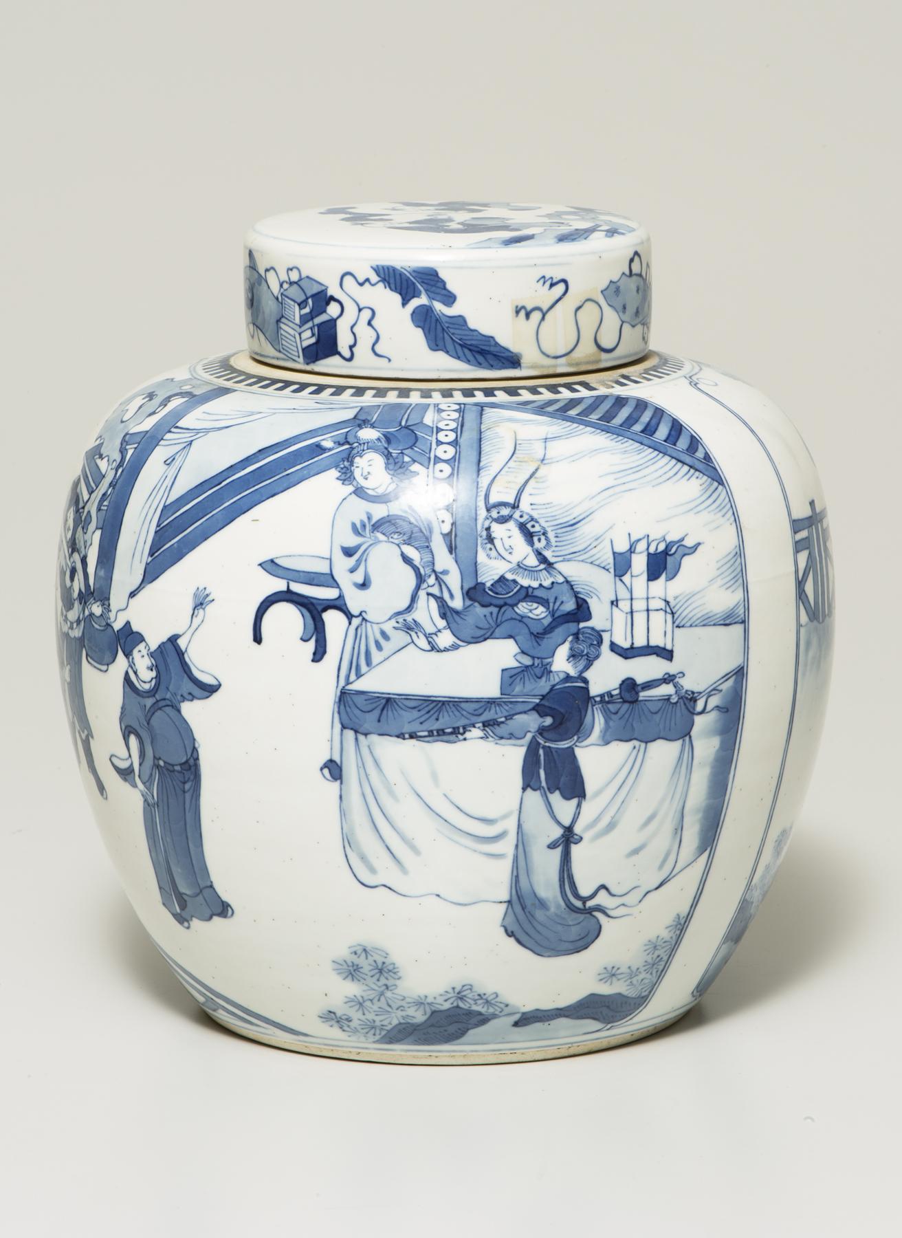 a white lidded jar is decorated in blue with two figues standing on a wooden balcony