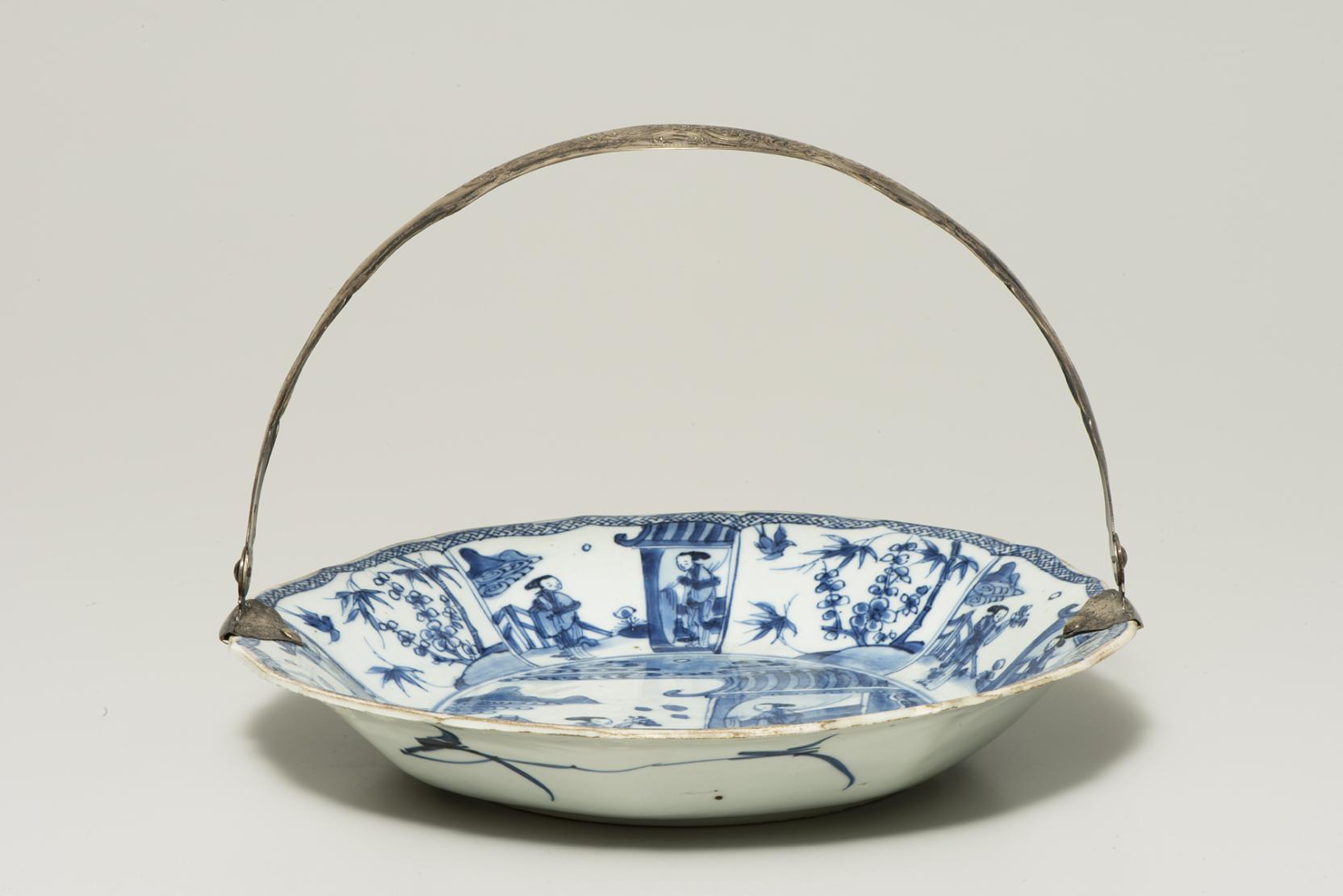 A large white dish with a tarnished silver handle is decorated with a variety of scenes, all in blue; at center, one figure outside a house presents flowers to another standing inside