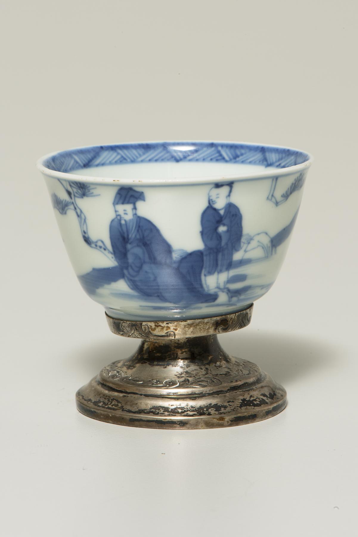 a small white cup with a silver foot is decorated in blue with an older man seated on the left and a younger man standing on the right