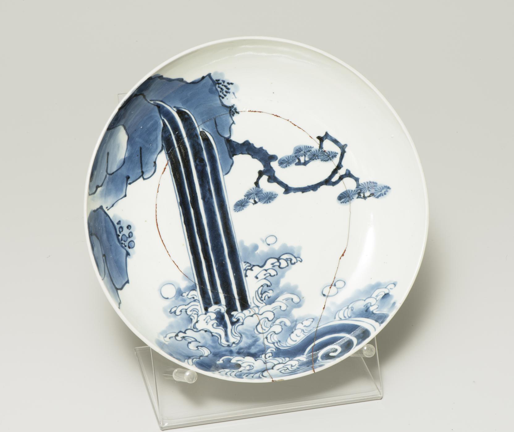 white plate with a blue scene: a waterfall crashes from rocks into water in front of a tree branch