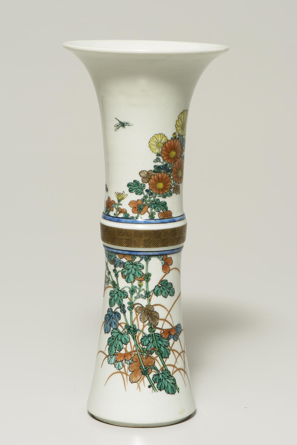 tall white vase with wide rim decorated with a brown band across the center; yellow and orange flowers with green leaves and stems emerge from the band in both directions