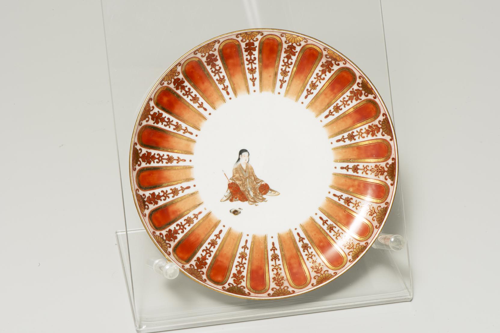 white plate with a seated figure dressed in red and gold at center, bordered by a repeating red arch and swirled design alternating pattern