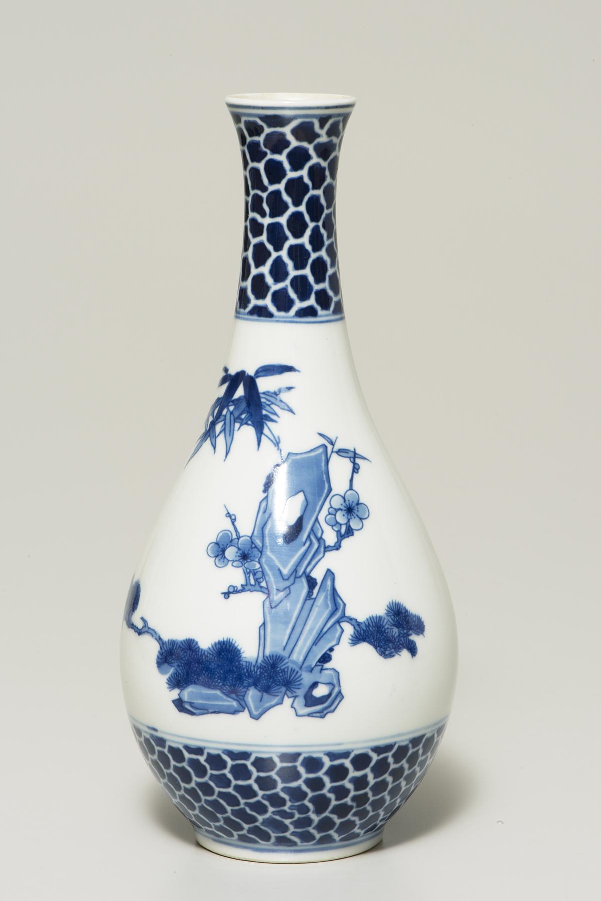A bulbous white vase with a long, narrow neck is decorated with bamboo, pines and rocks planted in blue; a dark blue geometric pattern covers the base and the neck 