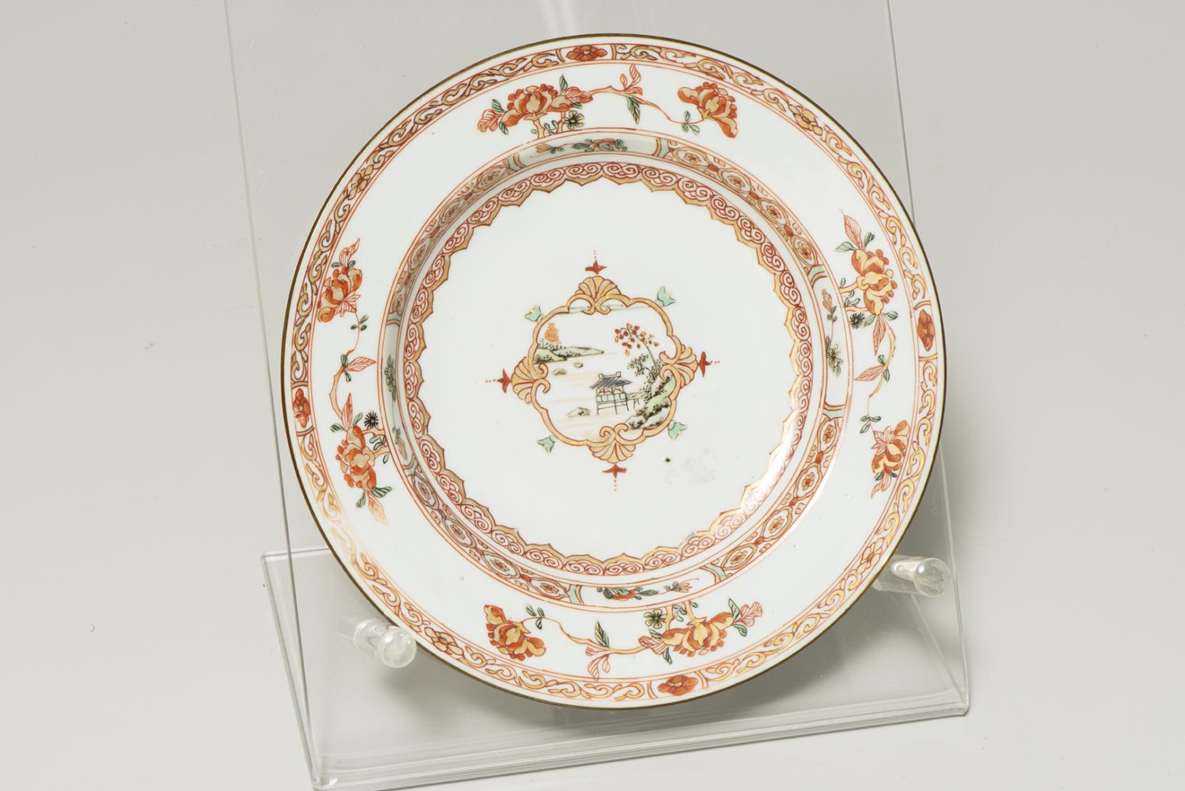 white plate decorated with a small outdoor scene at center, surrounded by a swirling red-orange pattern, then four evenly spaced orange flowers 